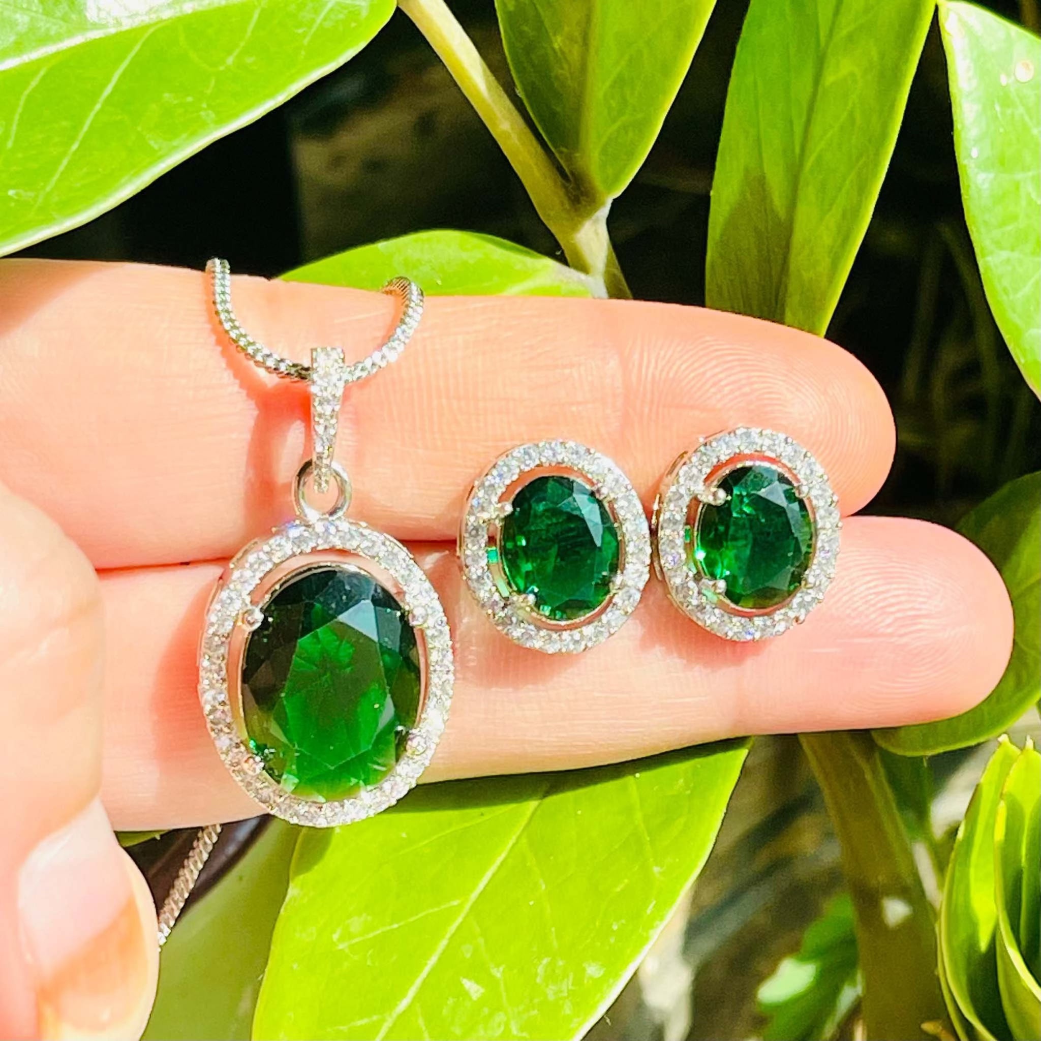 Emerald grand pendant set with oval green stone surrounded by white diamonds on silver chain and matching earrings against green leaves background