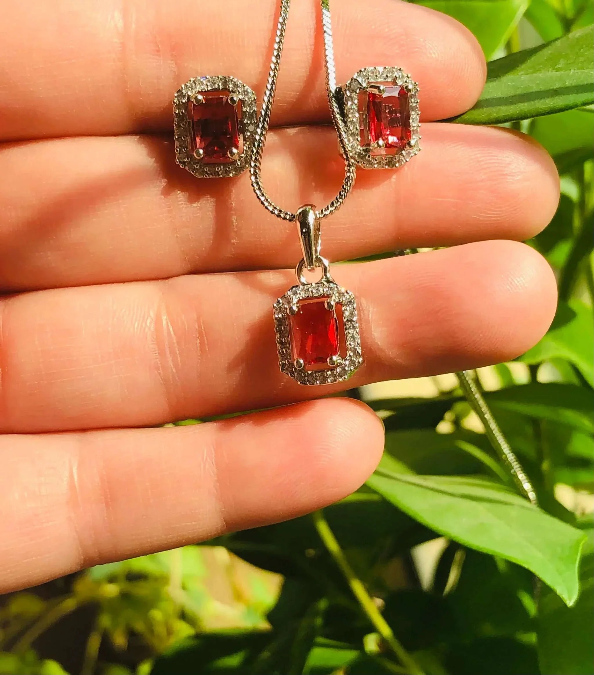 Red and silver rectangular pendant set with red gemstone and silver chain held against green leaves background