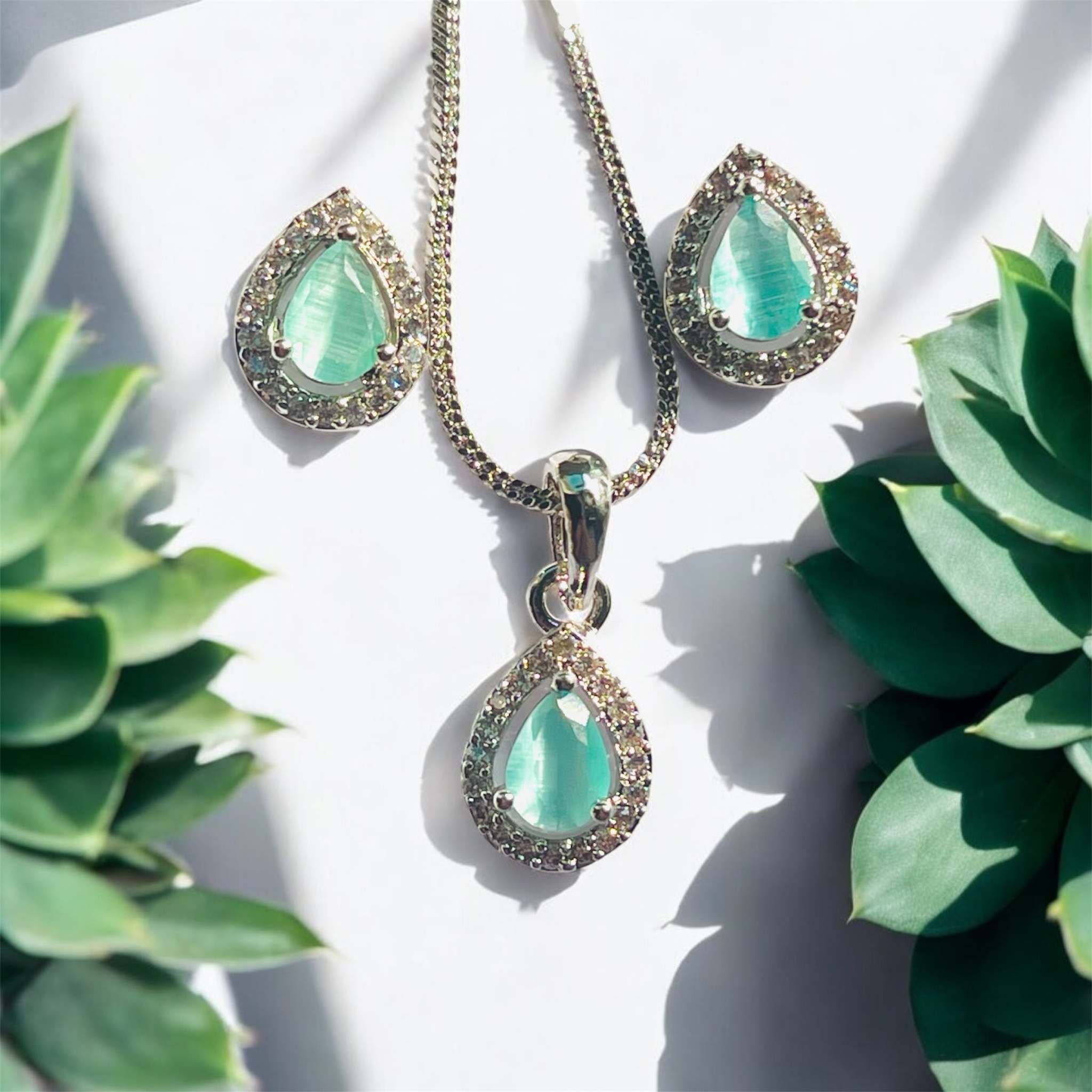 Teal and silver teardrop dainty pendant set with gemstone and chain on white background surrounded by green succulent plants