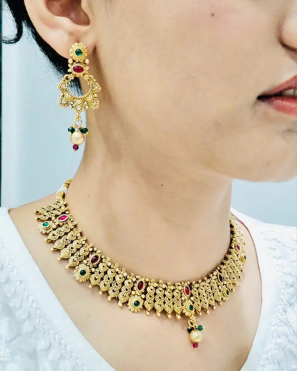 Woman wearing gold necklace and earring set with red and green stones.